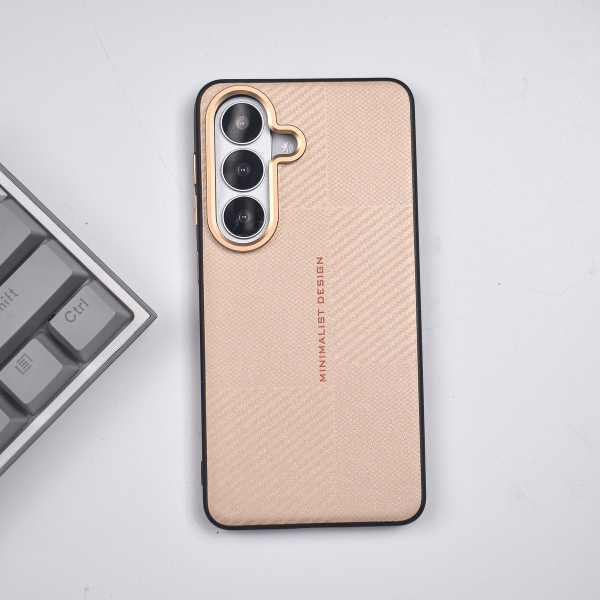 A minimalist phone case with a textured beige carbon-fiber pattern, black bumper edges, and a gold camera bezel, shown on a light gray surface next to a keyboard.
