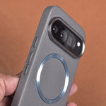 A close-up of a gray textured phone case with a metallic MagSafe ring, held by a hand against a warm brown background.