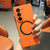Person holding an orange Samsung phone case in a casual setting with chairs in the background